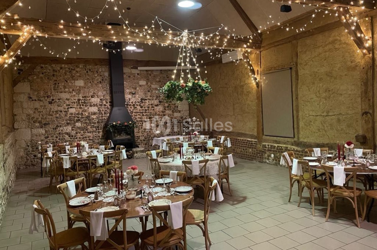 Salle de réception rustique avec tables rondes dressées, guirlandes lumineuses suspendues et murs en pierre apparente.