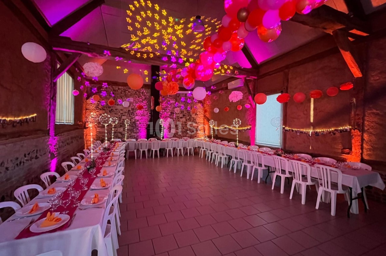 Salle décorée pour un événement, avec tables alignées, nappes rouges, ballons suspendus et éclairage coloré.