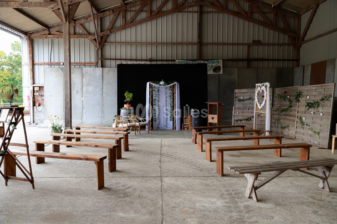 Salle de cérémonie rustique avec bancs en bois, décor floral et arche ornée de tissu dans un hangar ouvert.