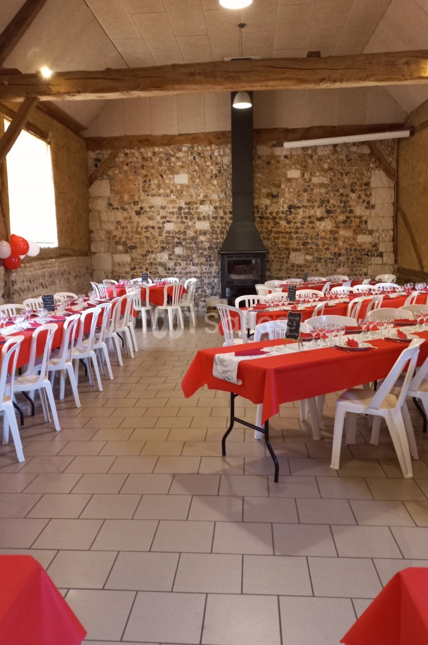 Salle décorée pour un événement, avec tables rouges dressées, chaises blanches et un poêle noir au fond.
