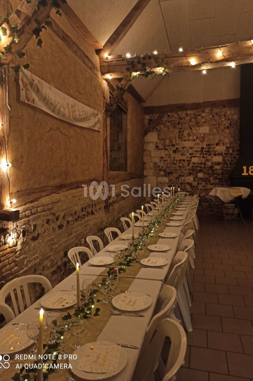 Grande table décorée pour un dîner, avec bougies et guirlandes lumineuses, dans une salle en pierres apparentes.