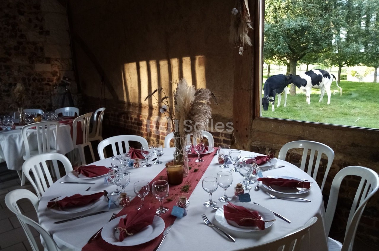Salle de réception rustique avec table dressée et vue sur des vaches dans un pré à travers une grande fenêtre.