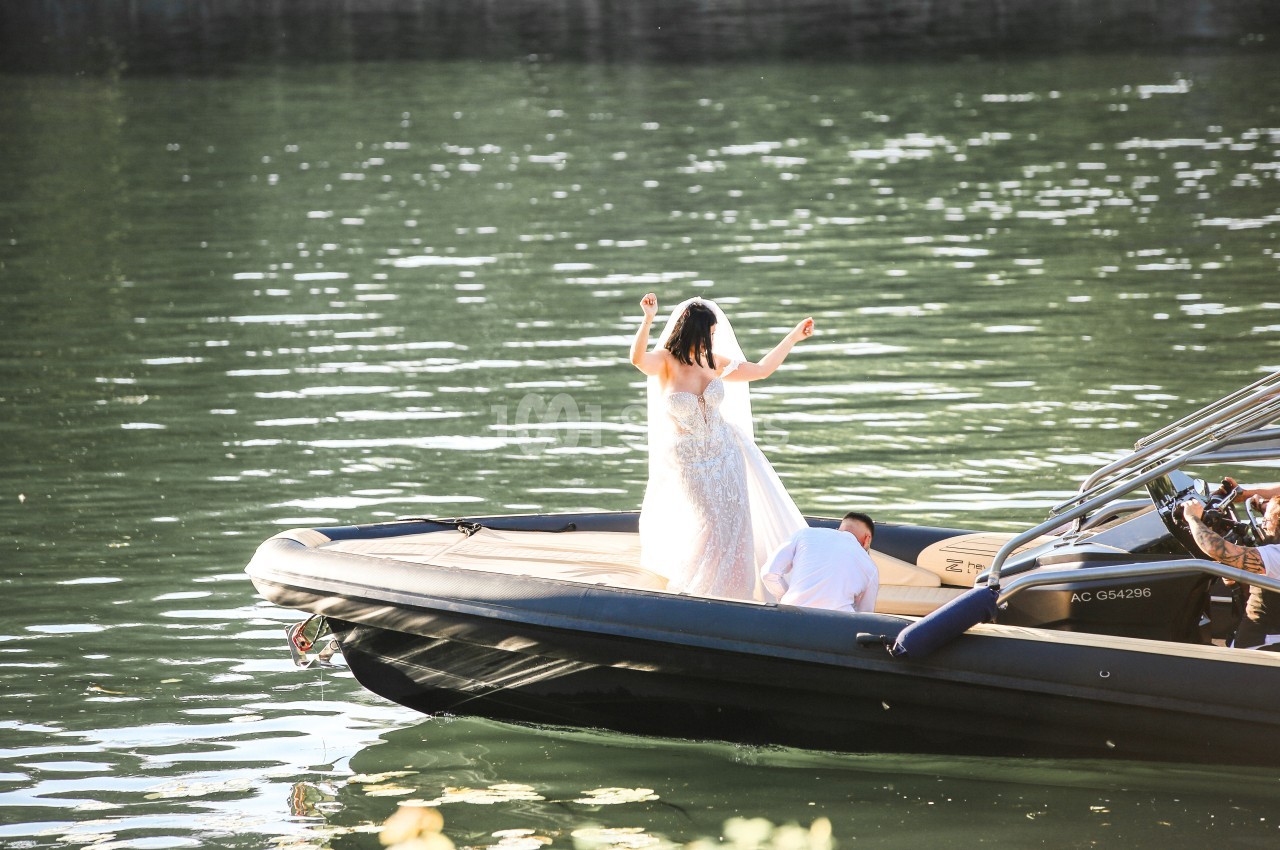 Une mariée en robe blanche se tient debout sur un bateau noir naviguant sur une eau calme et ensoleillée.
