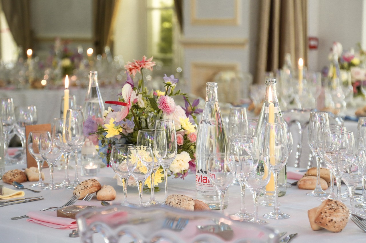 Table élégamment dressée avec des fleurs colorées, des verres, des bougies et des bouteilles d'eau dans une salle lumineuse.