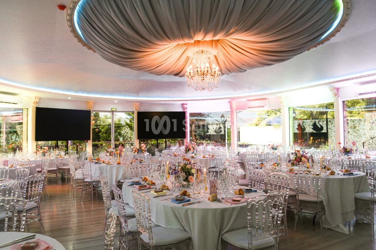 Salle de réception élégante avec tables dressées, chaises transparentes et grand lustre sous un plafond drapé.