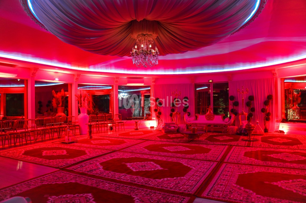 Salle de réception décorée de tapis rouges, éclairée par des lumières roses et un lustre central.