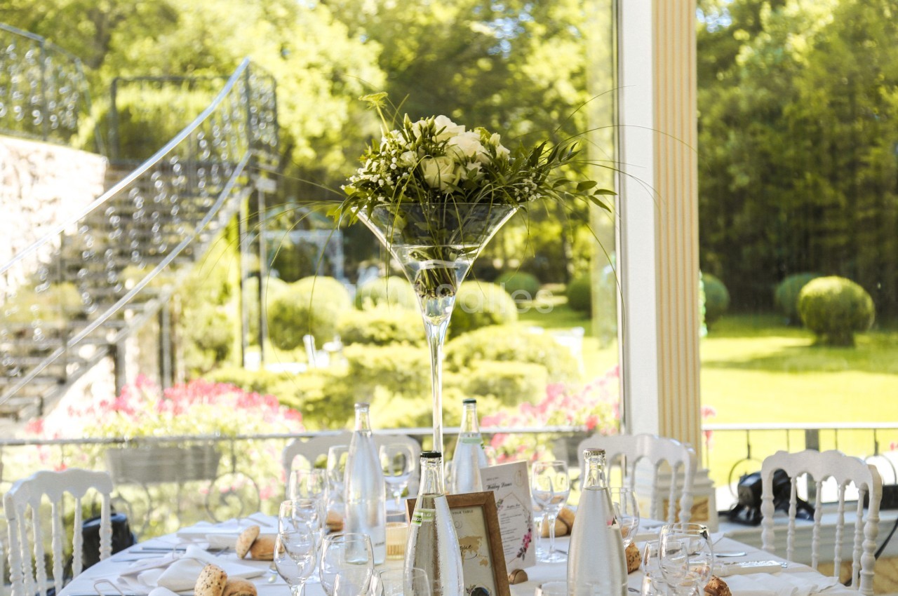 Centre de table floral sur une table dressée, avec vue sur un jardin verdoyant à travers de grandes baies vitrées.