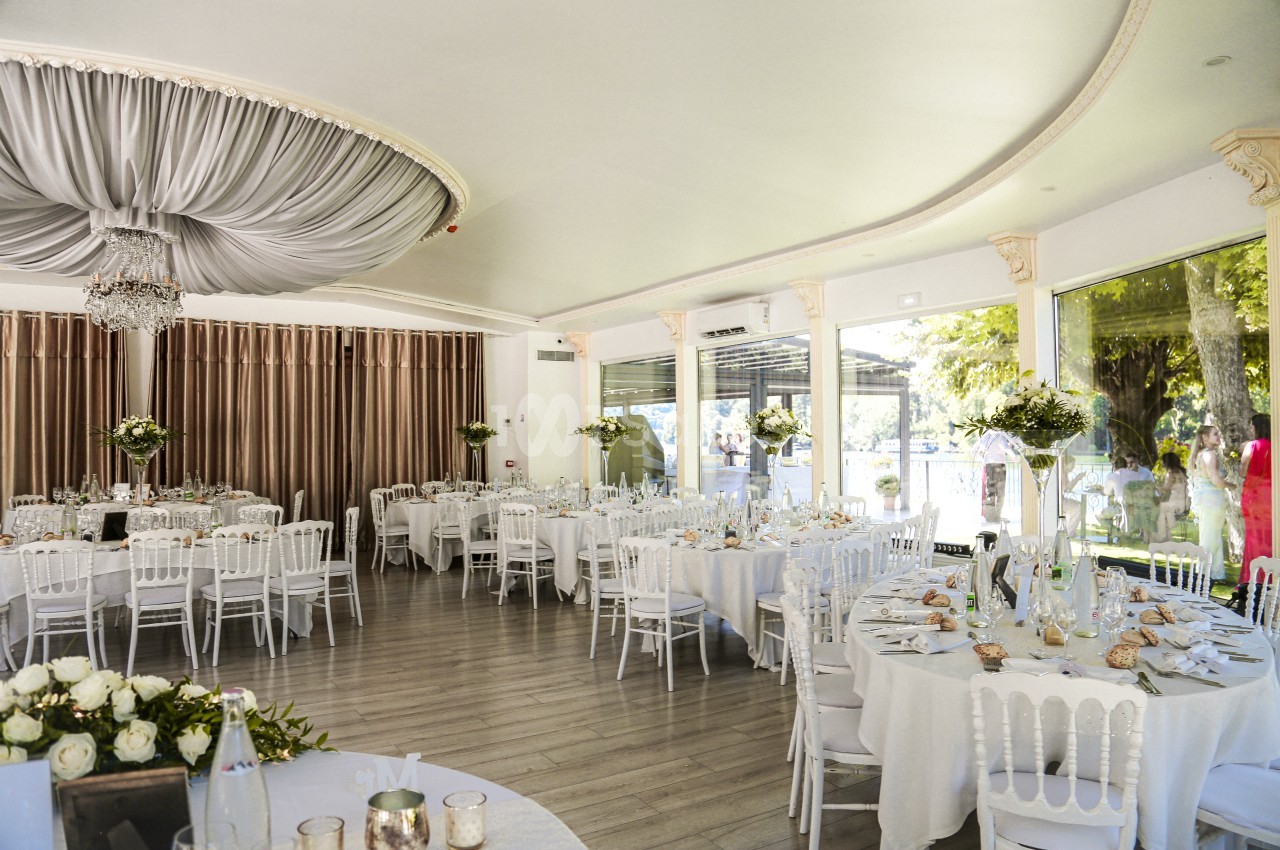 Salle de réception lumineuse avec tables rondes décorées, chaises blanches et grandes baies vitrées donnant sur un jardin.