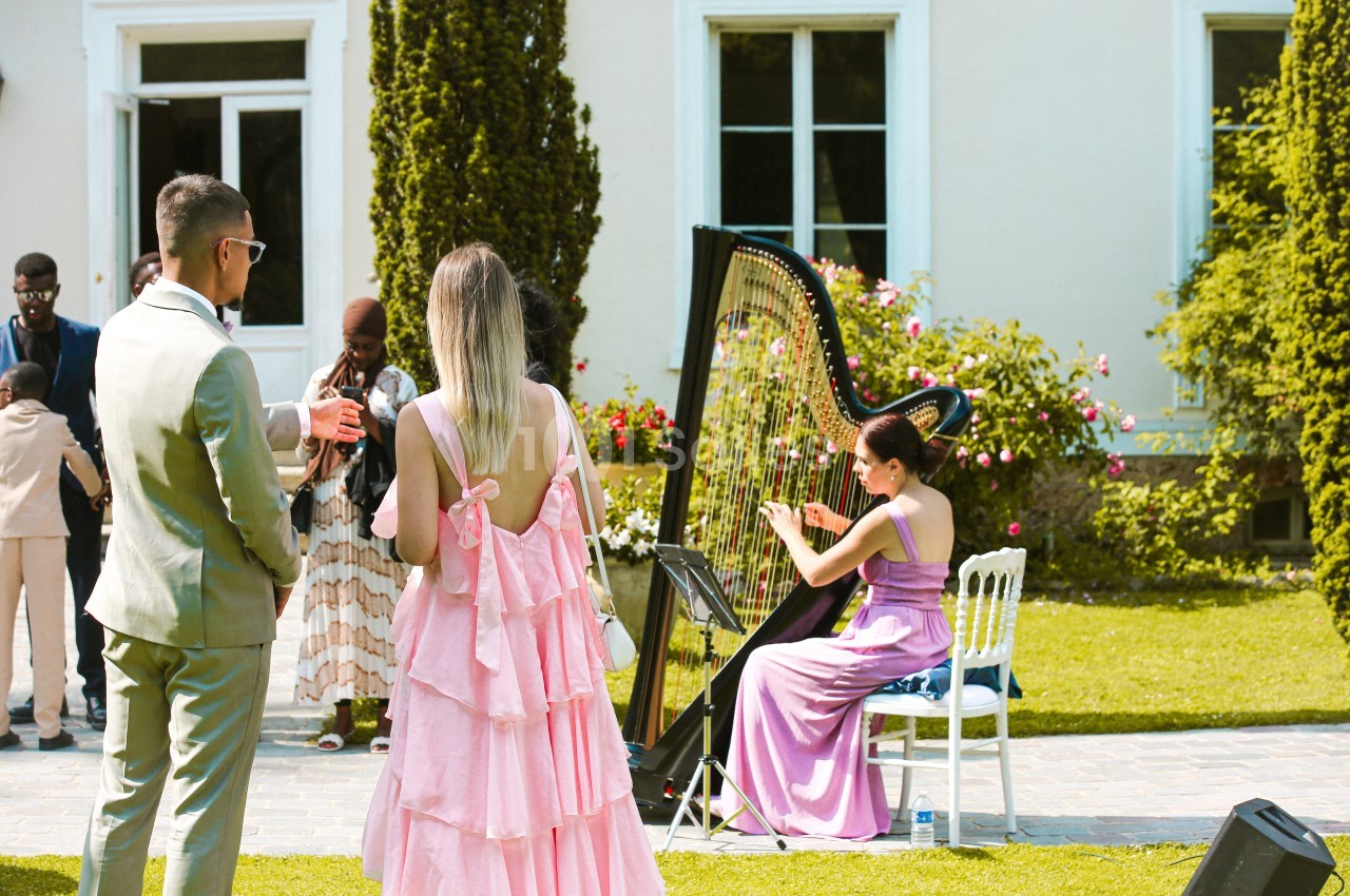Une harpiste joue en plein air lors d'un événement, entourée d'invités élégamment vêtus dans un jardin.