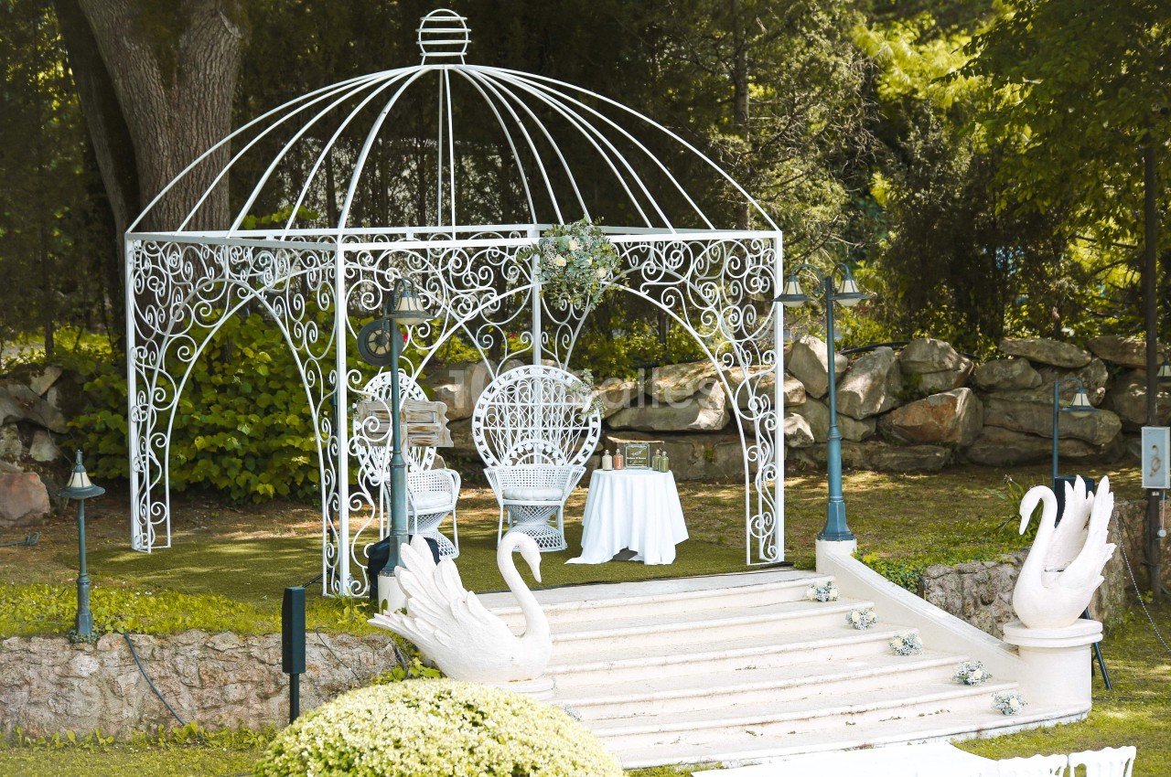 Kiosque en fer forgé blanc avec chaises et table, entouré de verdure et décoré de sculptures de cygnes.