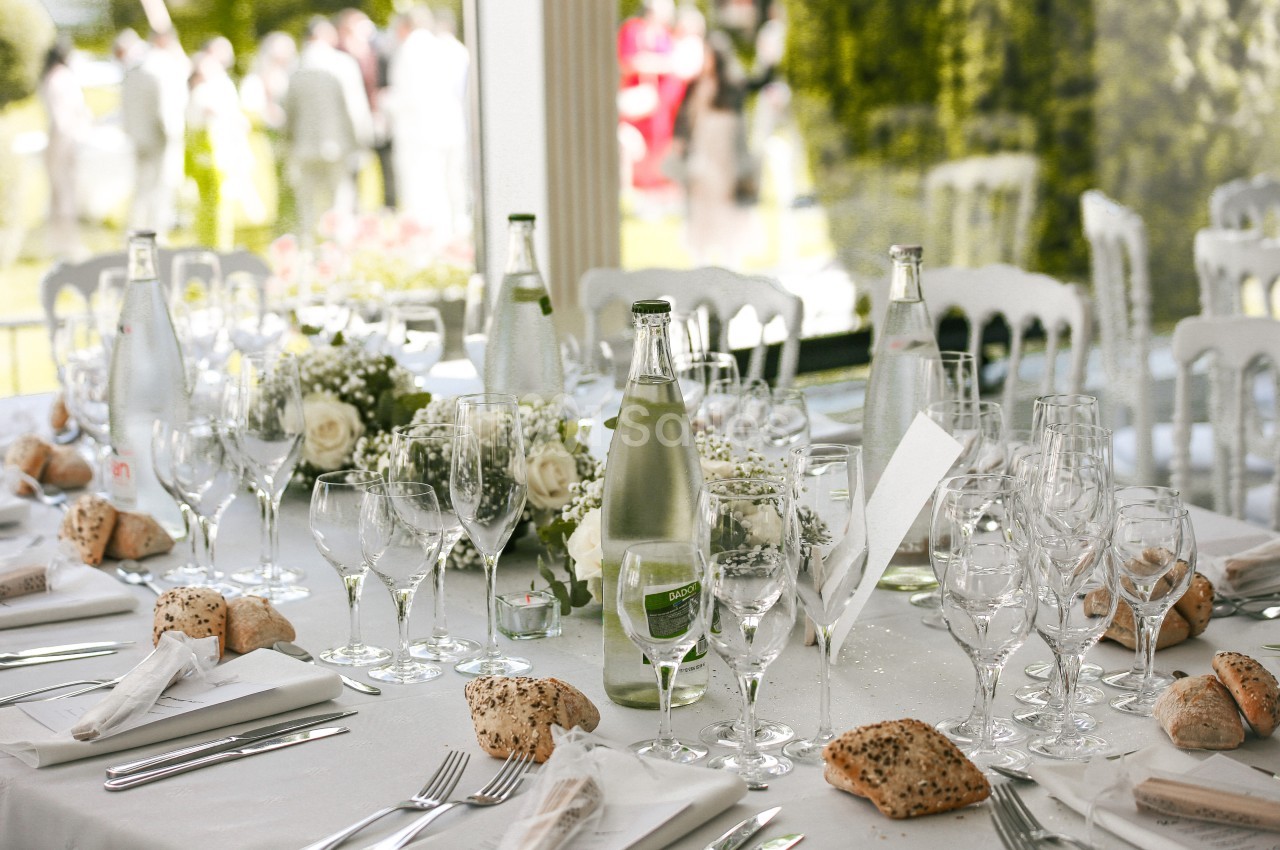 Table élégamment dressée avec nappes blanches, verres, bouteilles d'eau, fleurs et petits pains, en extérieur.