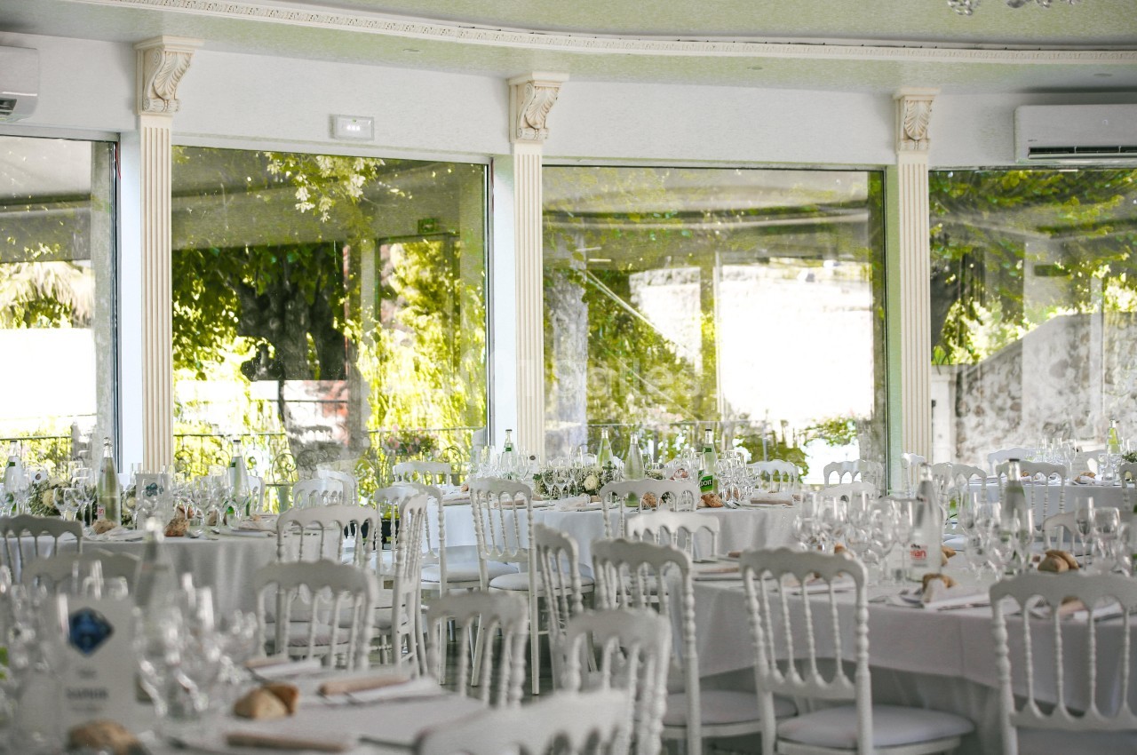 Salle de réception lumineuse avec tables rondes dressées, chaises blanches et grandes baies vitrées donnant sur un jardin.