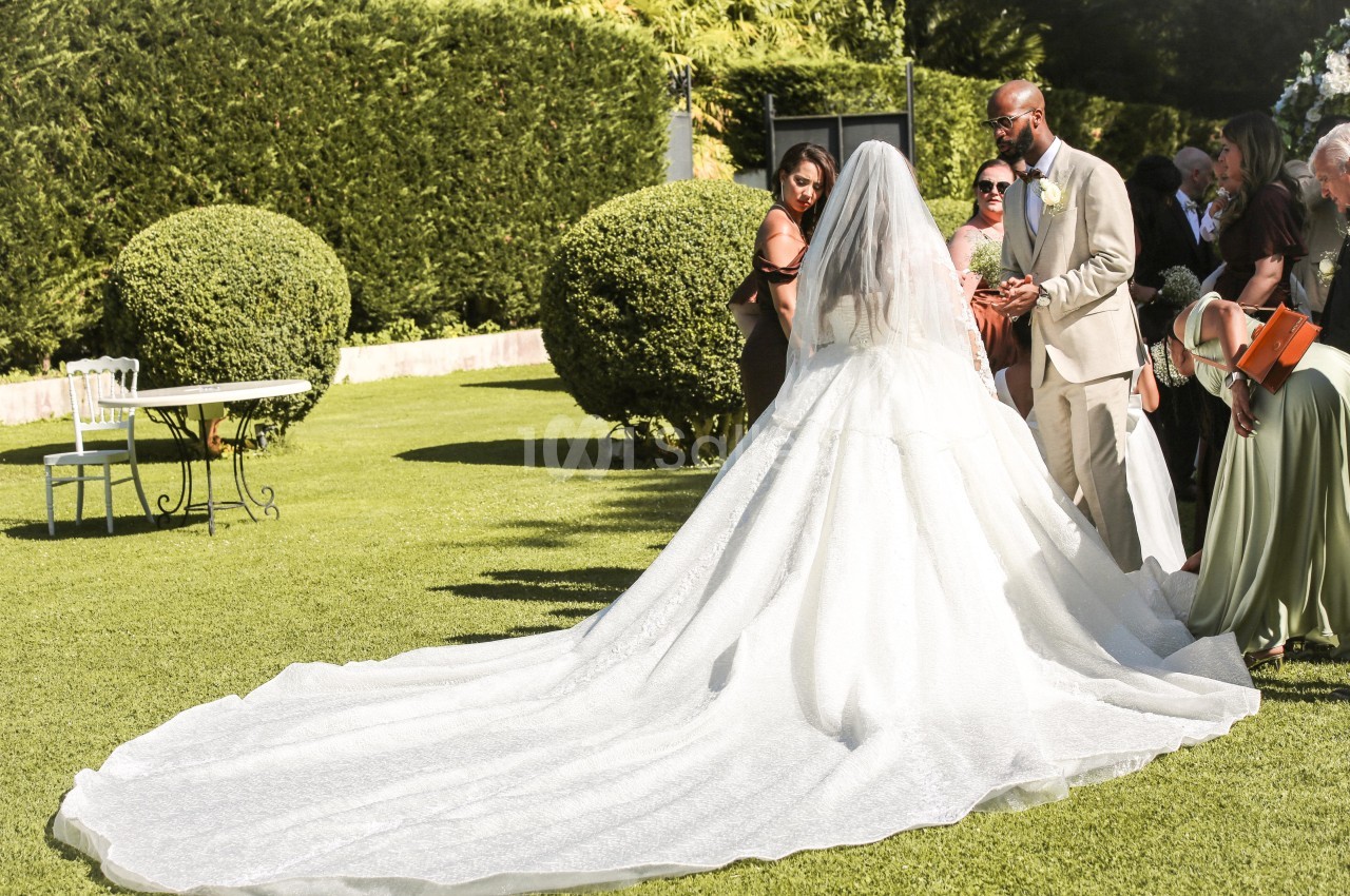 Une mariée en robe blanche avec une longue traîne se tient dans un jardin entourée d'invités.