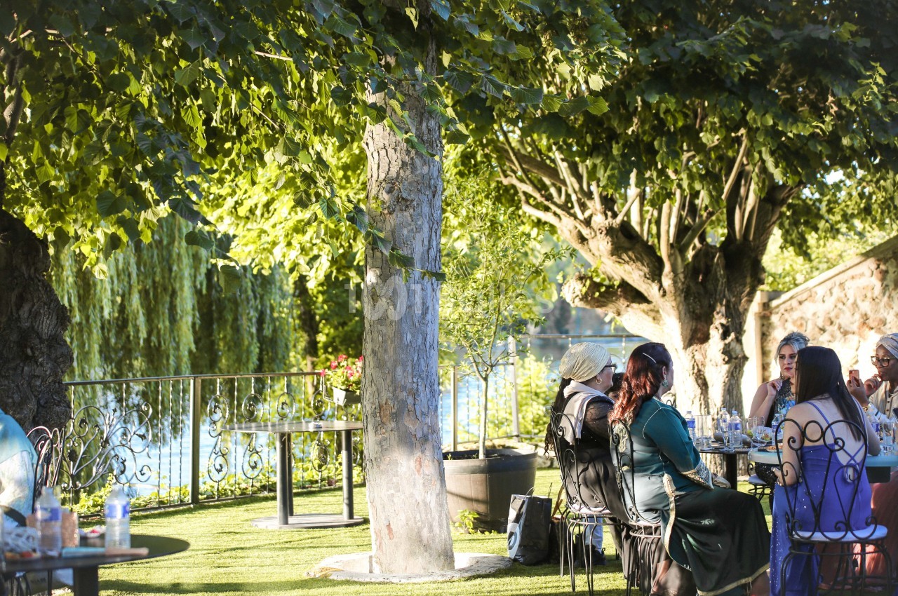 Des personnes assises à des tables dans un jardin ombragé près d'un plan d'eau, entourées d'arbres et de verdure.