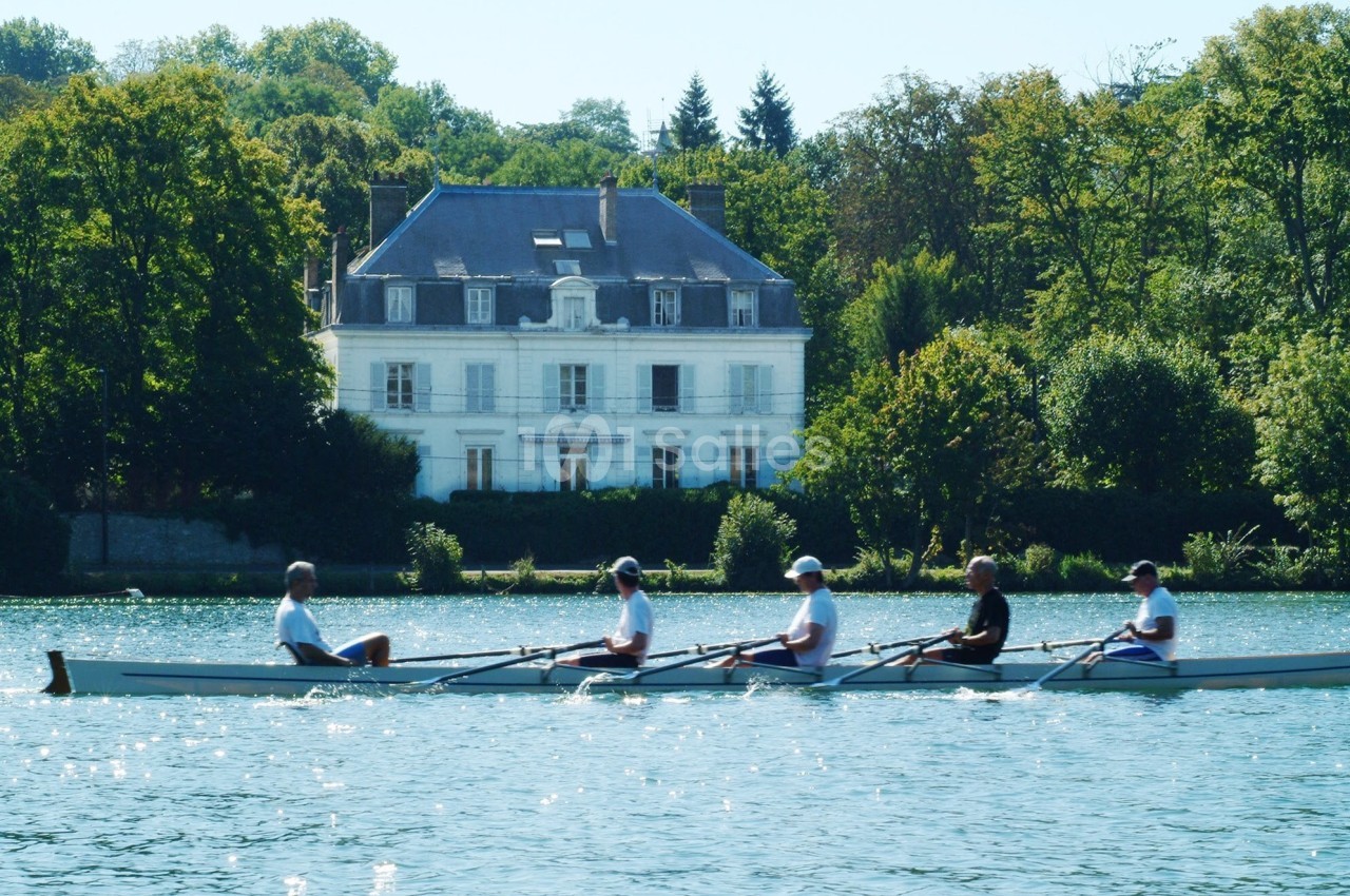 Quatre personnes rament sur un cours d'eau calme, avec un grand bâtiment blanc entouré d'arbres en arrière-plan.