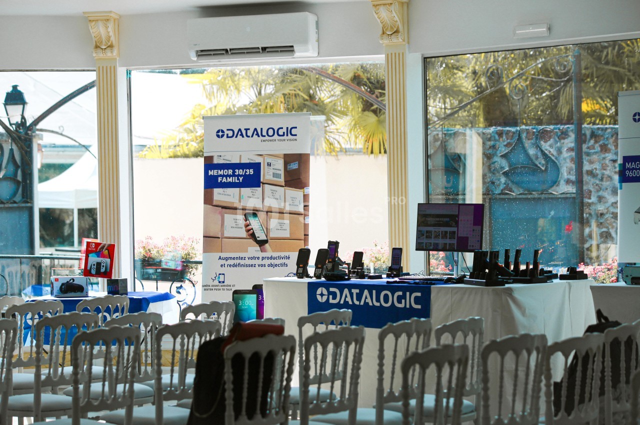 Salle équipée de chaises blanches, avec des stands présentant des produits technologiques et des affiches promotionnelles.