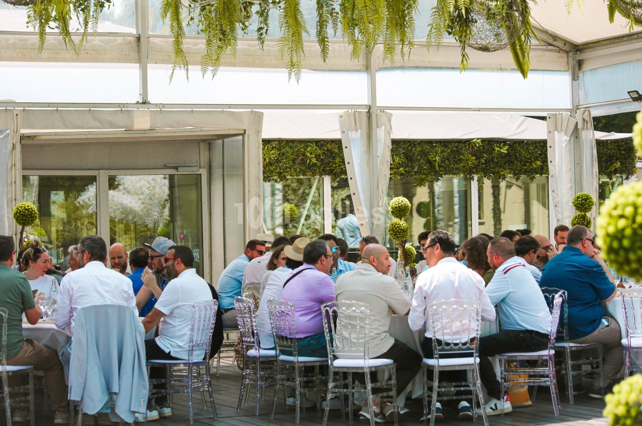 Groupe de personnes assises autour de tables dans un espace extérieur couvert, décoré de plantes suspendues.