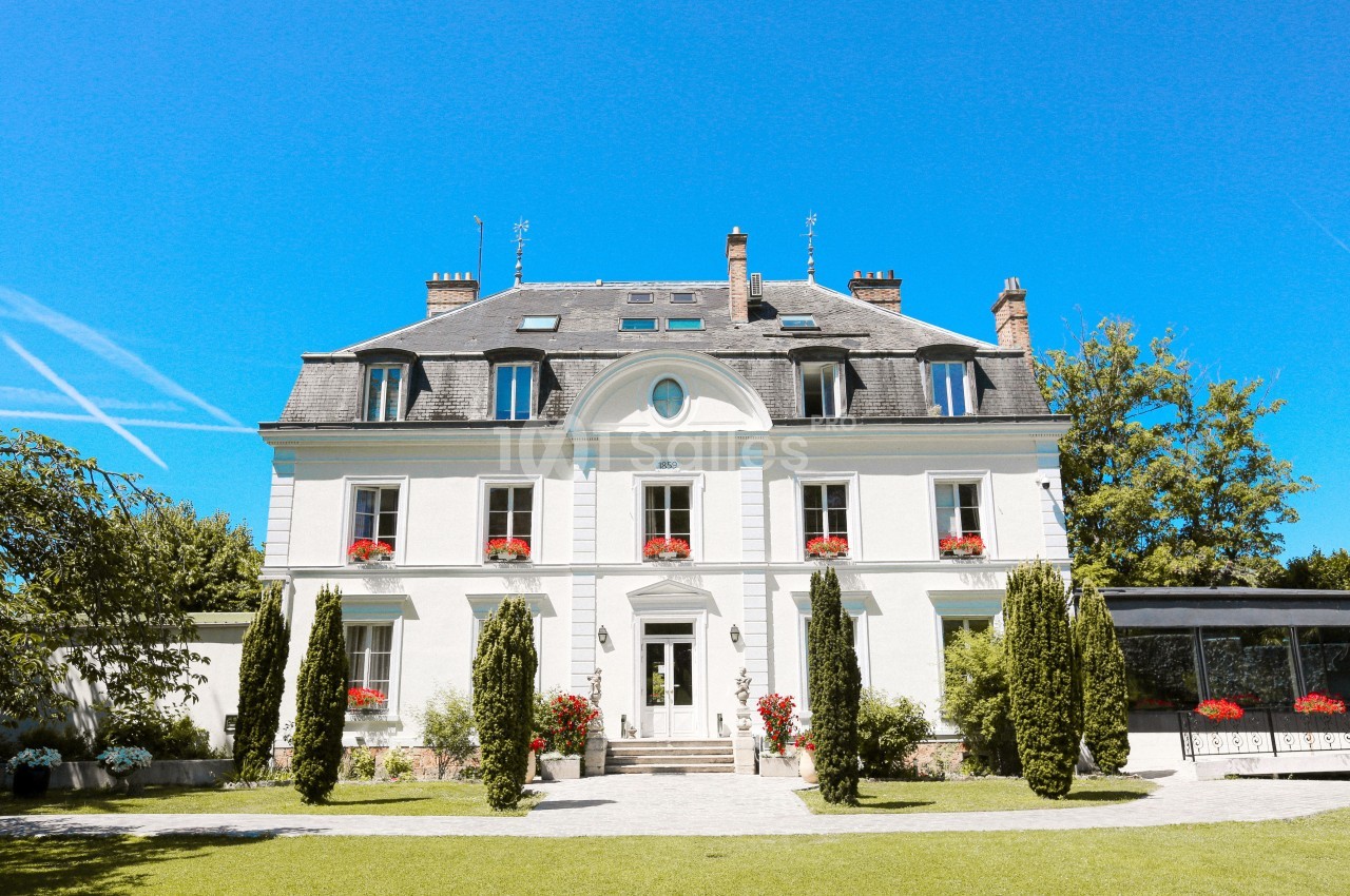Façade d'une grande maison blanche avec des volets bleus, entourée d'arbres et d'un jardin sous un ciel bleu.