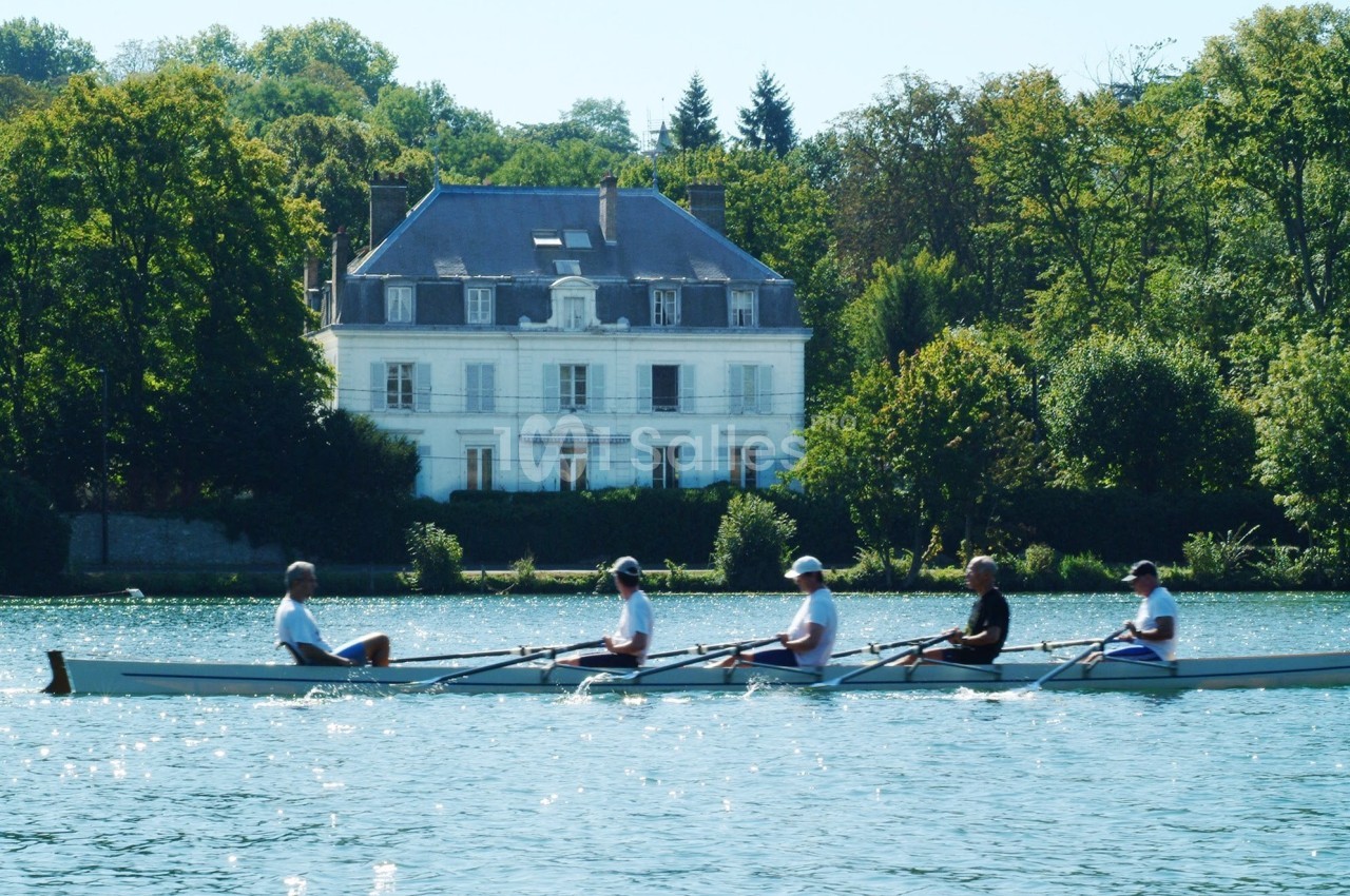 Quatre personnes rament sur une rivière devant une grande maison blanche entourée d'arbres.