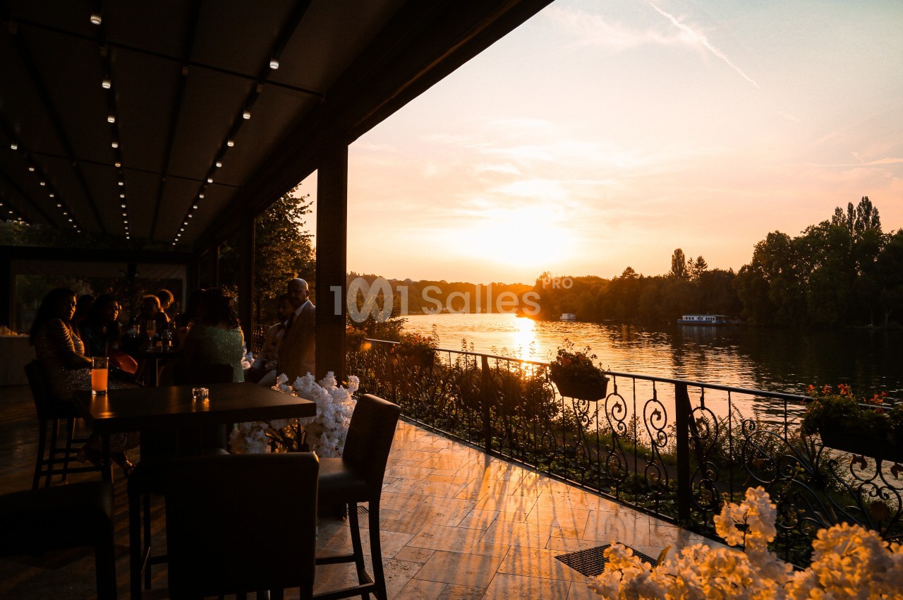 Terrasse au bord d'une rivière au coucher du soleil, avec des tables, des chaises et des fleurs en premier plan.