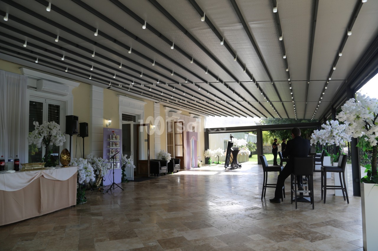 Terrasse couverte avec éclairage suspendu, tables, chaises et décorations florales, donnant sur un jardin verdoyant.