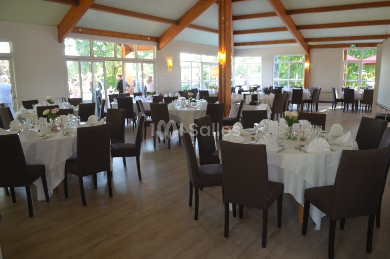 Salle de réception lumineuse avec tables rondes dressées, nappes blanches et chaises sombres, vue sur l'extérieur.