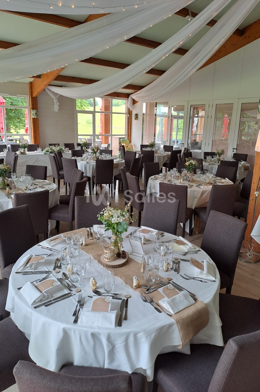 Salle de réception décorée avec des tables rondes dressées, nappes blanches, centres de table floraux et voilages suspendus.