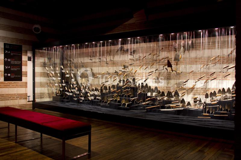 Vitrine exposant des outils anciens suspendus, éclairés dans un espace sombre avec un banc rouge au premier plan.