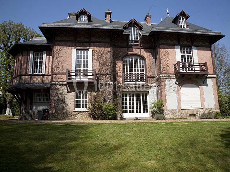 Grande maison en briques et pierre avec balcons, fenêtres à croisillons et jardin verdoyant au premier plan.