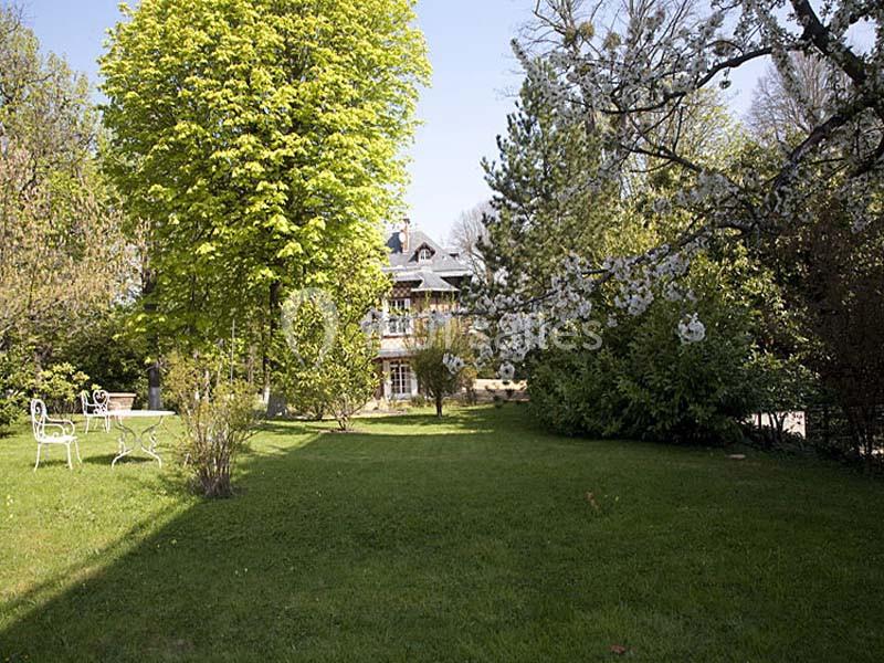 Jardin verdoyant avec pelouse, arbres en fleurs et mobilier de jardin blanc devant une maison en arrière-plan.