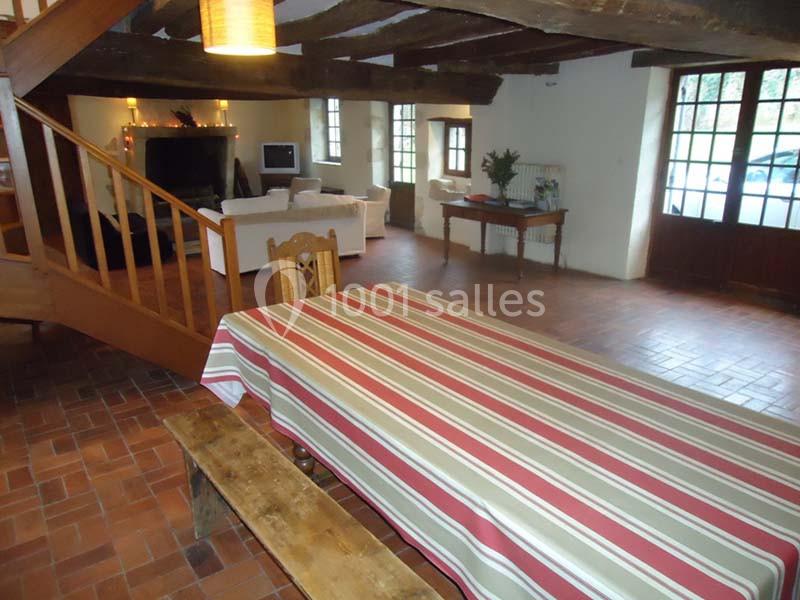 Salle à manger rustique avec table à rayures, banc en bois, escalier en bois et salon avec cheminée en arrière-plan.