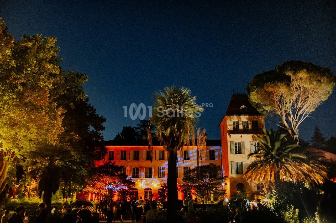 Façade d'un bâtiment éclairé de lumières colorées, entouré d'arbres et de végétation, en soirée.