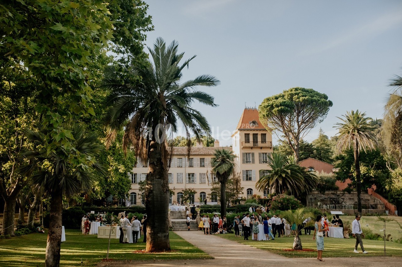 Jardin avec palmiers et pelouse, devant un grand bâtiment ancien, où des personnes sont rassemblées lors d'un événement.