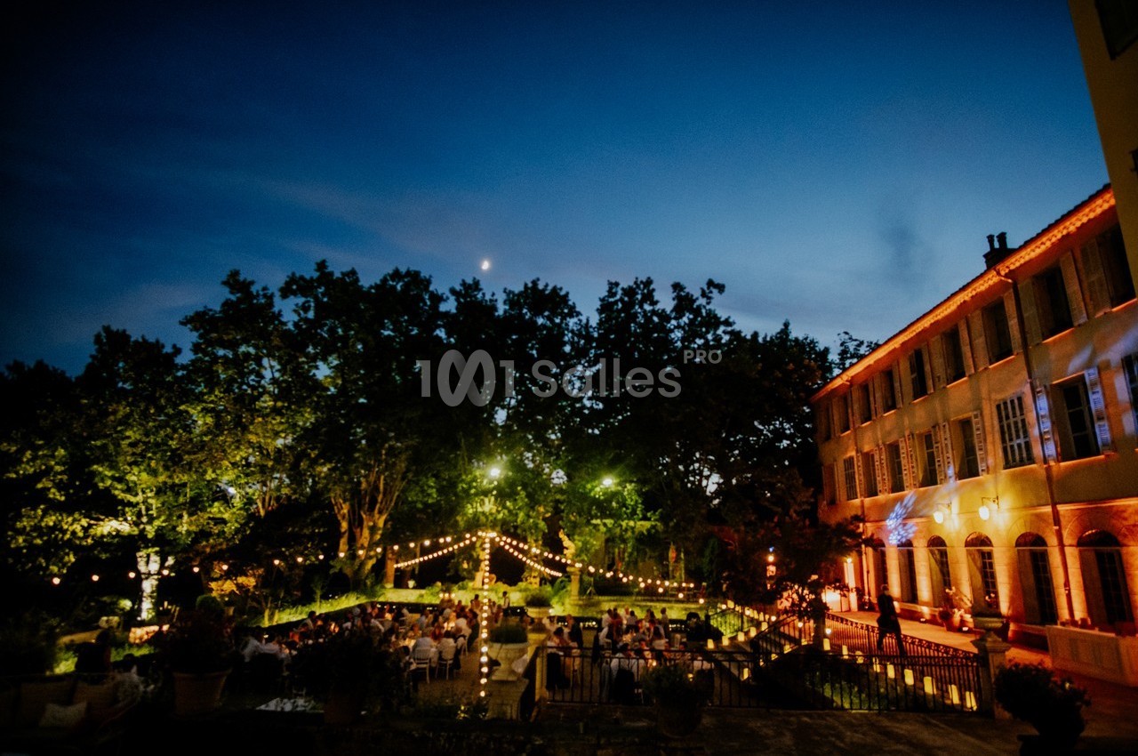 Jardin éclairé par des guirlandes lumineuses avec des invités assis, près d'un bâtiment illuminé au crépuscule.
