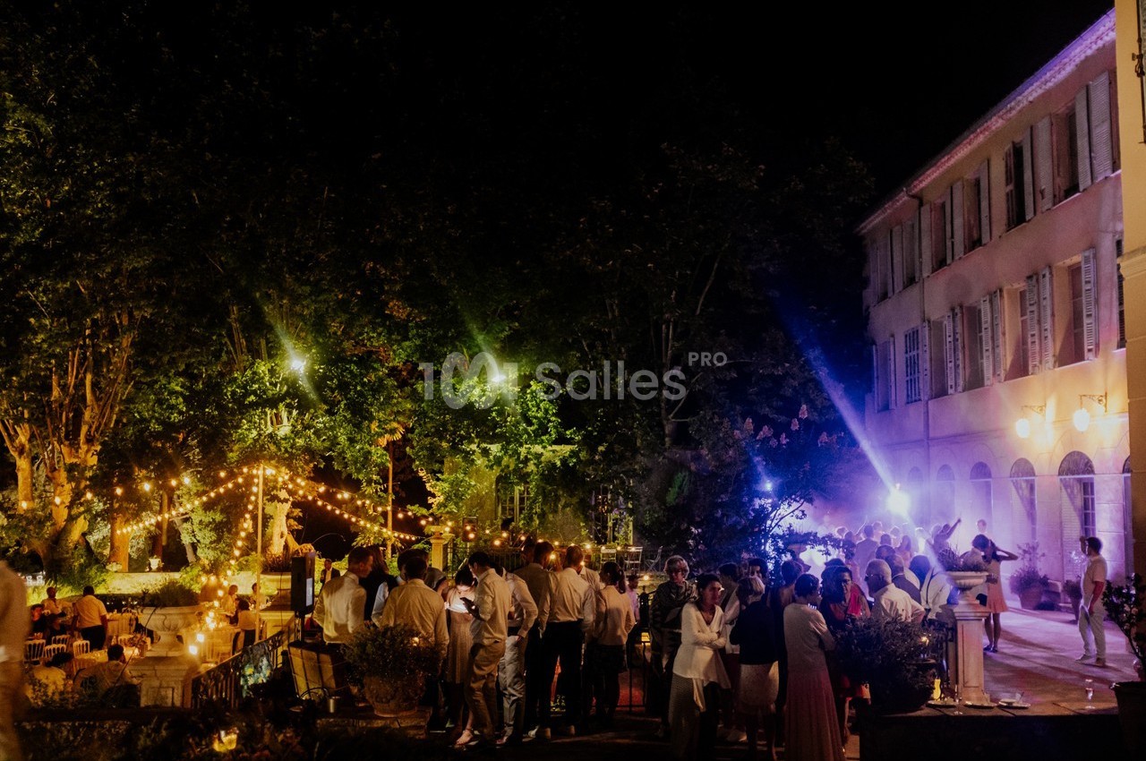 Groupe de personnes réunies en soirée dans un jardin éclairé par des guirlandes lumineuses près d'un bâtiment ancien.