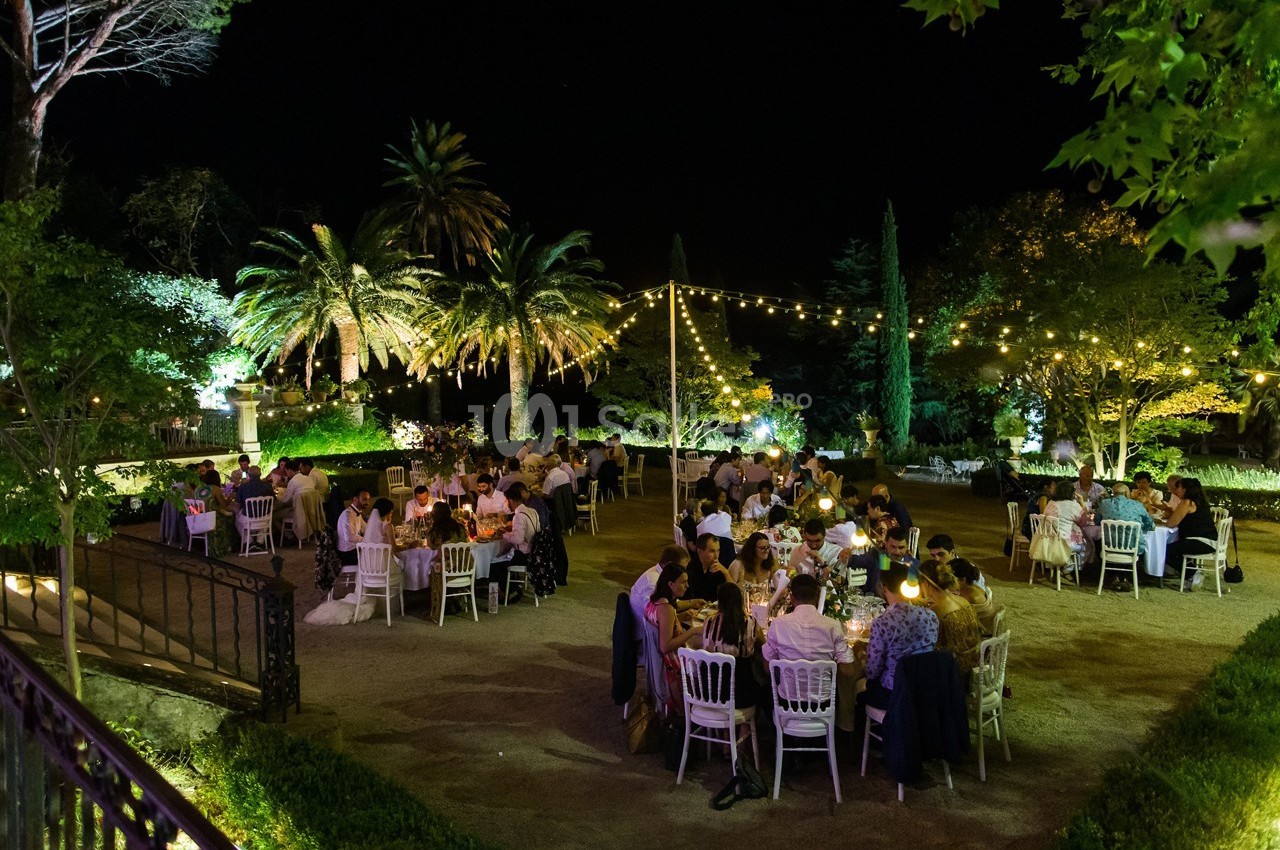Des convives dînent en plein air dans un jardin éclairé par des guirlandes lumineuses, entouré de palmiers et de verdure.