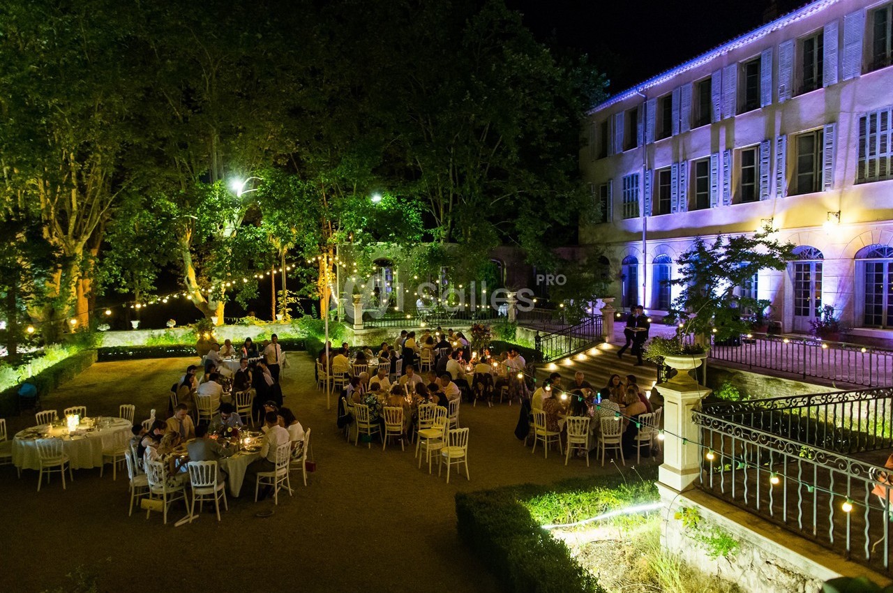Personnes dînant en plein air dans un jardin éclairé, près d'un bâtiment ancien illuminé de nuit.