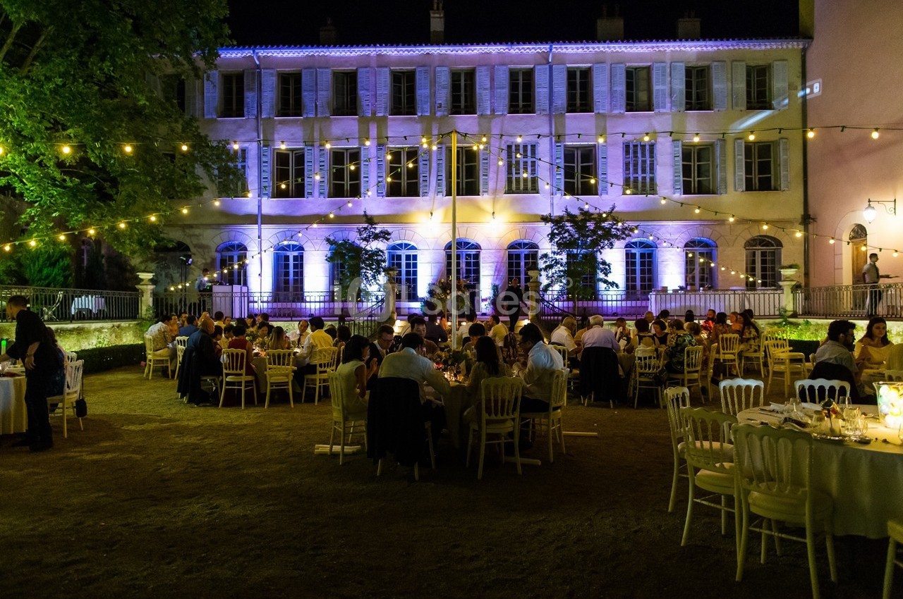 Dîner en plein air dans une cour éclairée par des guirlandes lumineuses, devant un bâtiment illuminé de nuit.