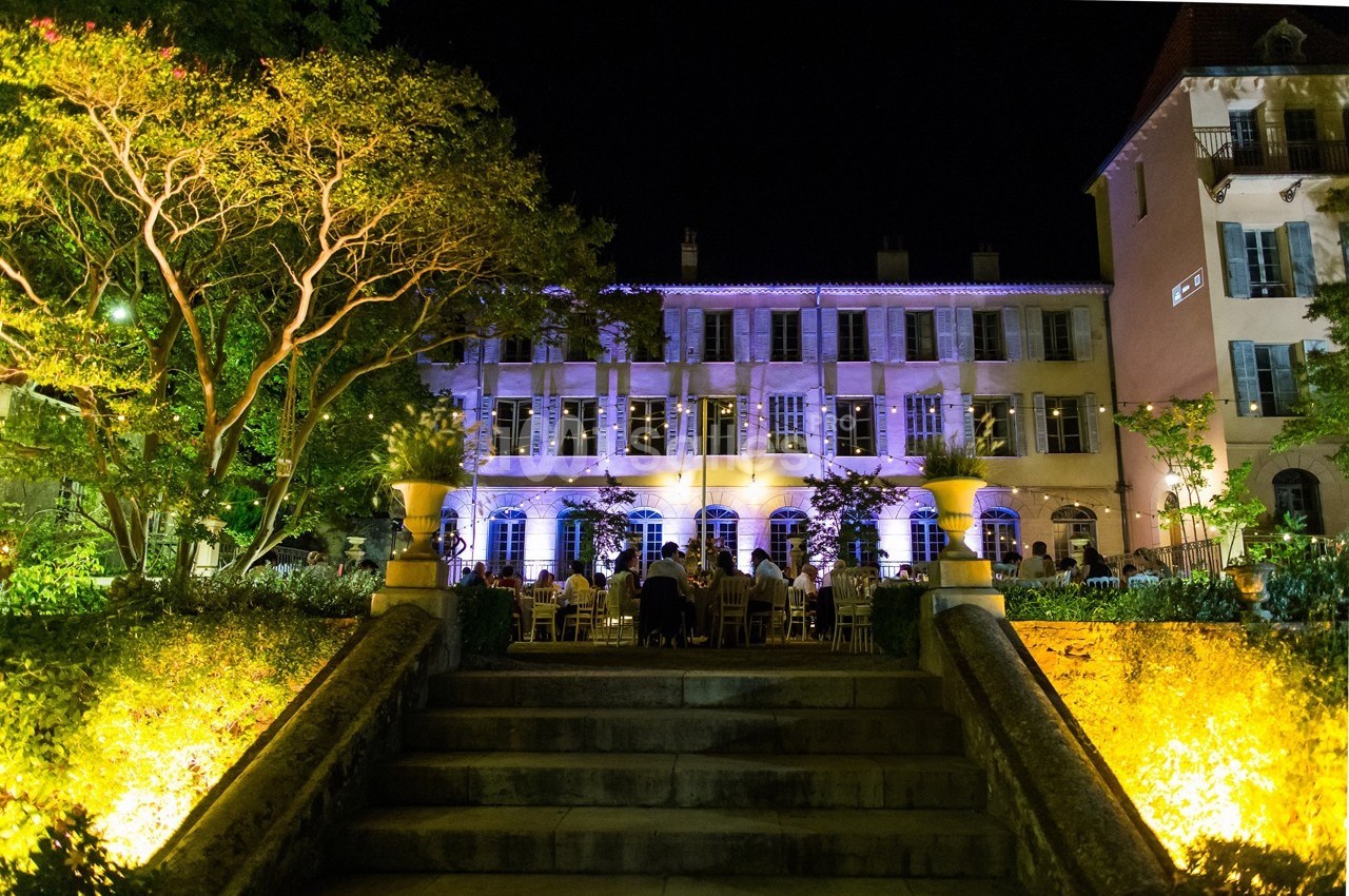 Façade d'un bâtiment éclairé la nuit, entouré de jardins avec des invités assis à des tables en extérieur.