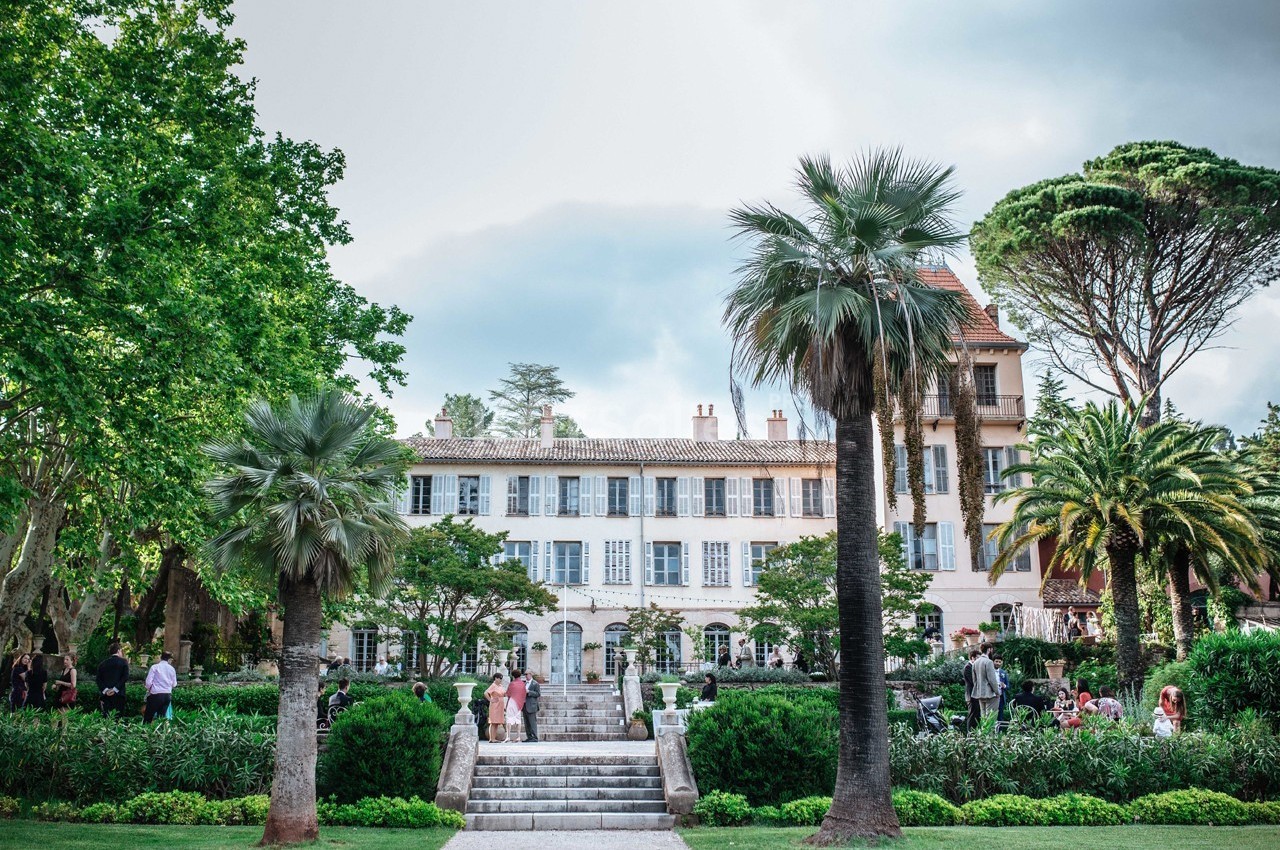 Façade d'une grande maison entourée de jardins verdoyants avec des palmiers et des personnes se promenant.