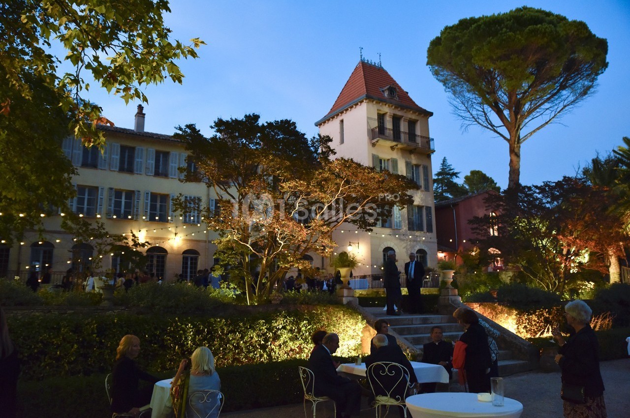 Façade d'une grande maison éclairée au crépuscule, avec des invités assis dans un jardin.