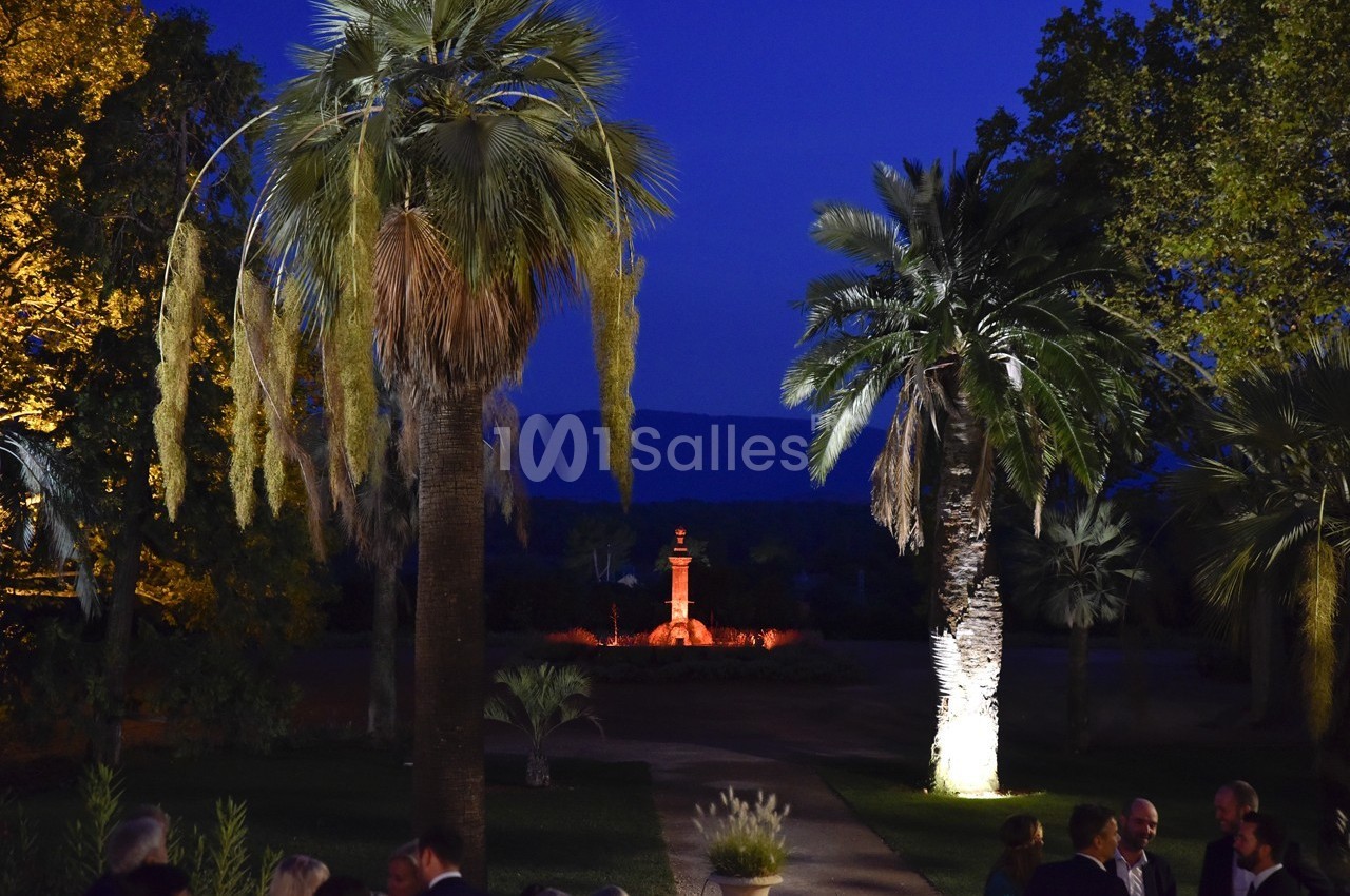 Allée bordée de palmiers éclairés, menant à une fontaine illuminée dans un jardin au crépuscule.