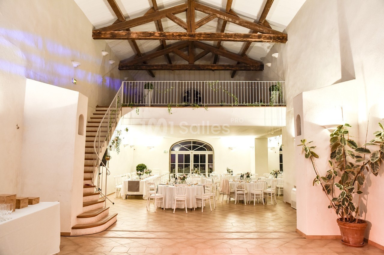 Salle de réception lumineuse avec tables dressées, escalier en colimaçon et poutres apparentes au plafond.