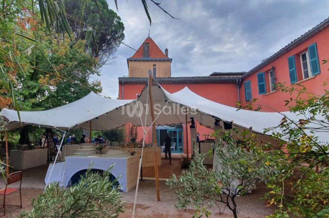 Tente blanche installée dans une cour extérieure devant un bâtiment rose à volets bleus, entouré de végétation.