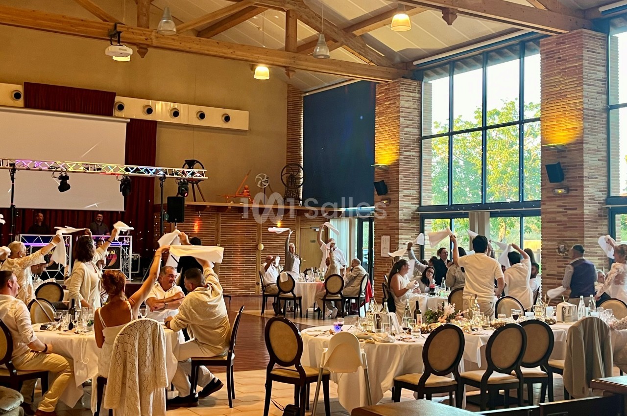 Salle de réception lumineuse avec des invités en tenue blanche célébrant autour de tables décorées.