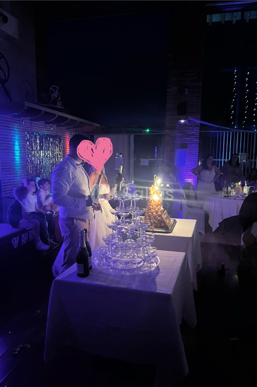 Un homme en tenue blanche près d'une fontaine de verres et d'une pièce montée éclairée dans une salle sombre.