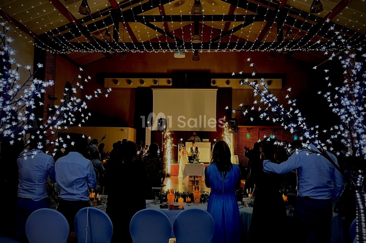 Salle de réception décorée avec des guirlandes lumineuses, des invités debout face à une scène éclairée.