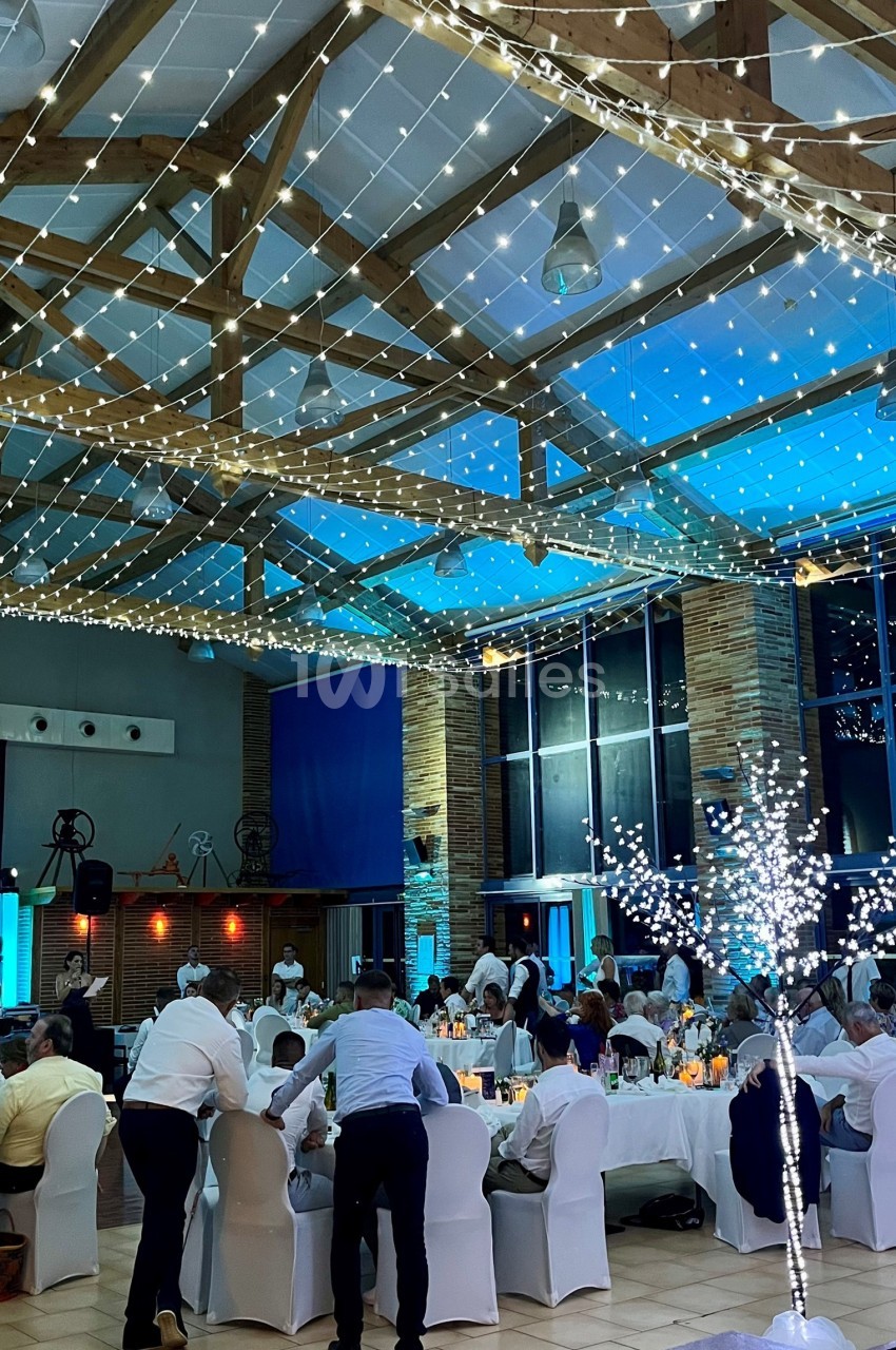 Salle de réception décorée avec des guirlandes lumineuses, des tables dressées et des invités en tenue de soirée.