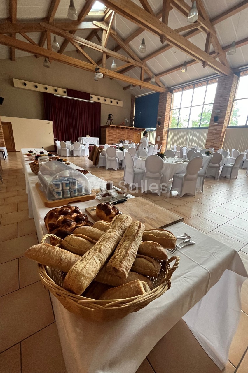 Salle de réception avec tables dressées et buffet comprenant des pains, viennoiseries et autres aliments.