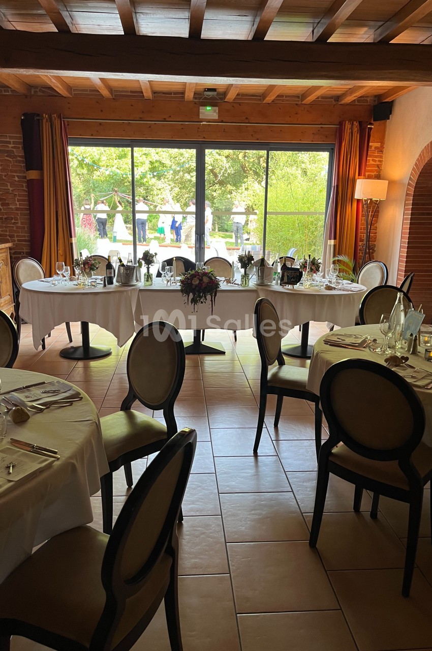 Salle de réception lumineuse avec tables dressées, chaises élégantes et vue sur un jardin à travers une baie vitrée.